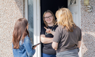 Three people are standing at the entrance of a house. One person is inside the house, partially visible through the open door, and two Settle colleagues are outside; one is holding a mobile phone and wearing glasses.
