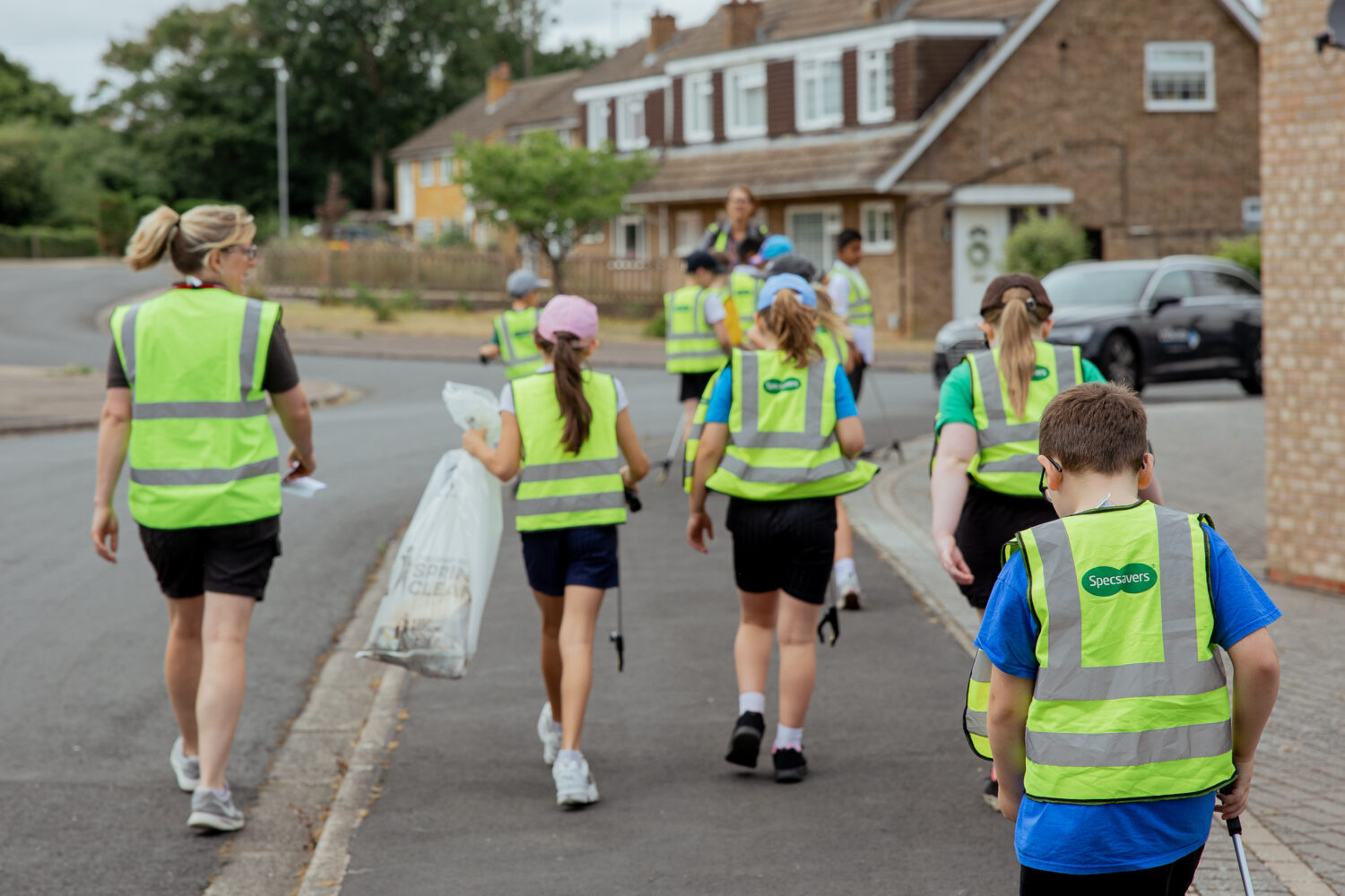 A group of school children and adults, including Settle colleagues walking along a residential street. They are wearing high-visibility vests and carrying litter picking equipment.