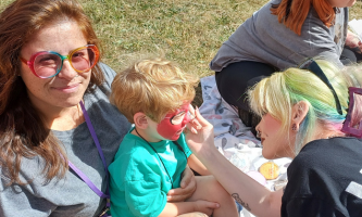 A group of people are sitting on a blanket on the grass. Councillor Sadie Billing, is holding her son. Another person with dyed hair is painting the boy's face while another woman and child are in the background.