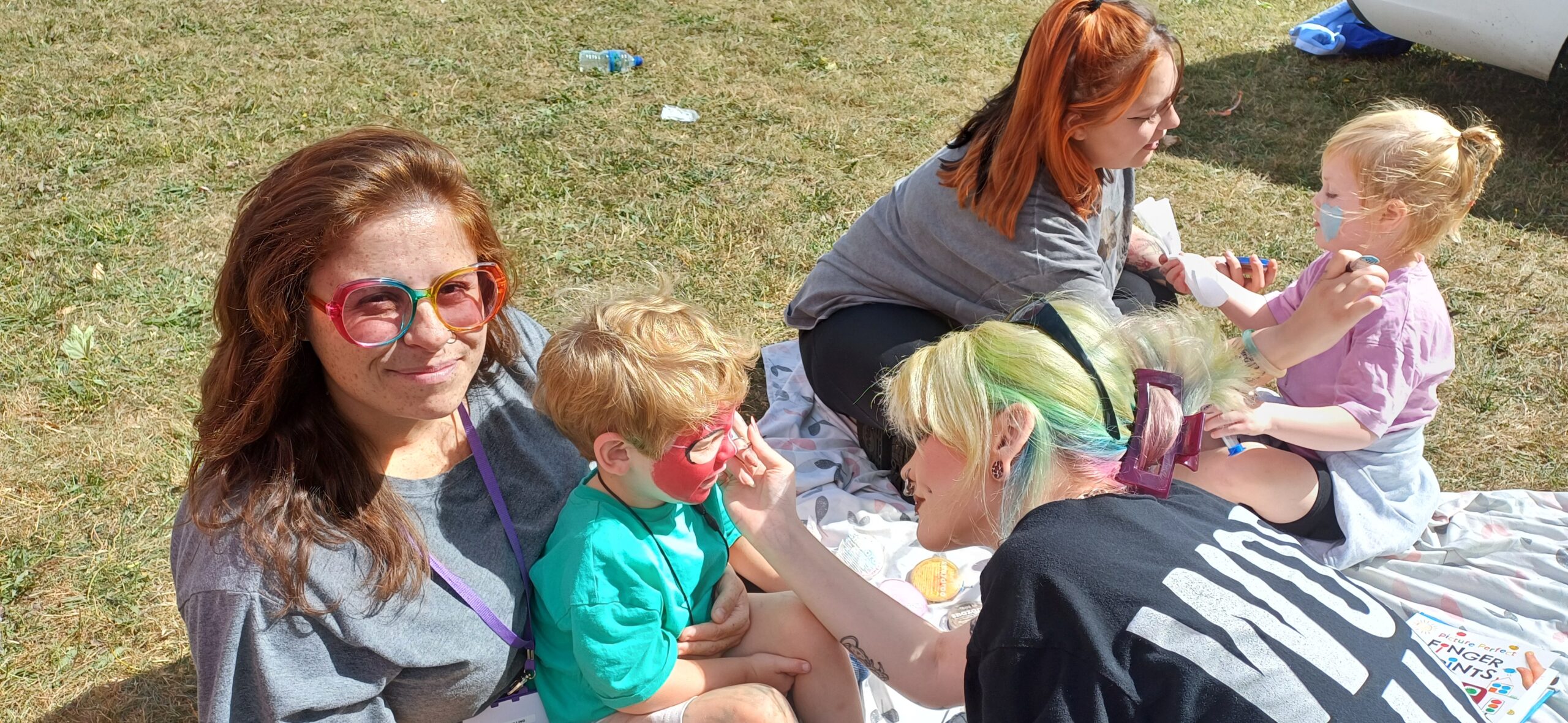 A group of people are sitting on a blanket on the grass at Jackmans Community Fun Day. Councillor Sadie Billing is holding her son while he has his face painted. 