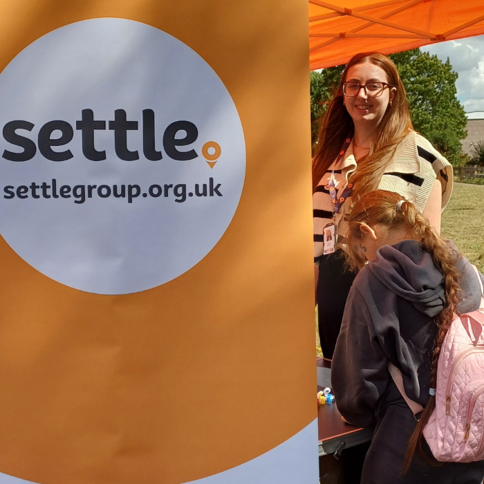 A Settle colleague stands under an orange tent beside a large orange banner displaying the Settle logo and website (settlegroup.org.uk). A child wearing a pink rucksack is in front of the banner engaging with activities on the stall. 