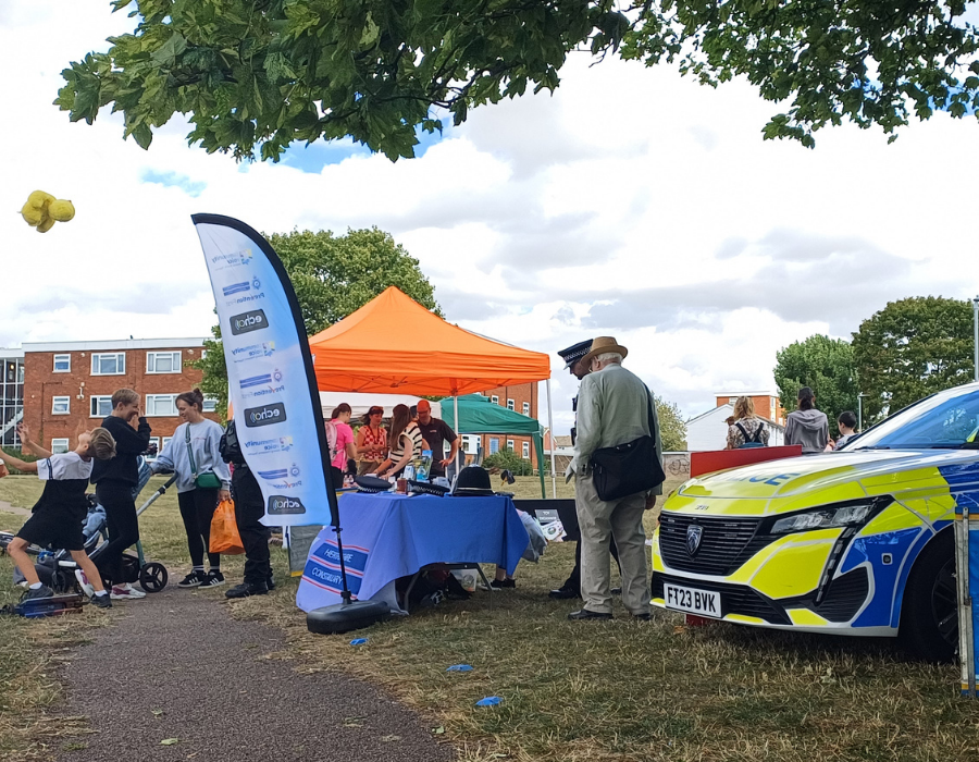 Jackmans community event with people gathered around stalls and activities. A blue table with a banner featuring the Herts Constabulary logo is in the foreground, an orange tent is in the background, and a police car is visible on the right. Residents and their families are interacting, playing, and riding scooters on the grass.