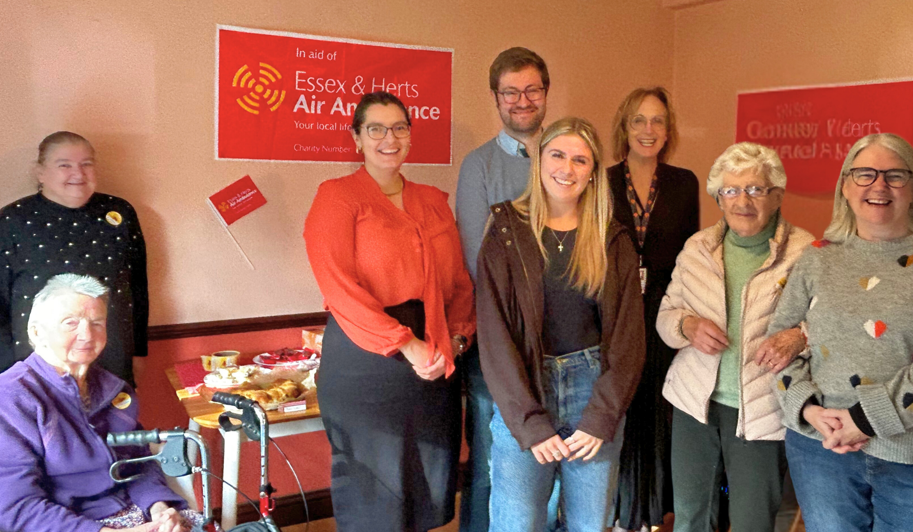 Settle colleagues with residents and councillors from North Herts Council at a charity cake sale. in front of two red banners that read “In aid of Essex & Herts Air Ambulance” and “Charity Event.” A table with plates of food and drinks is visible to the left, next to a seated person using a mobility aid. 