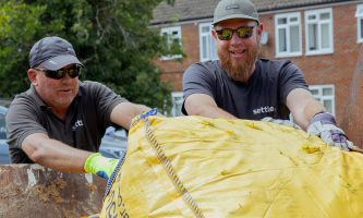 Two Settle colleagues wearing work gloves lift a large yellow bag filled with garden waste into a metal container, with green foliage in the foreground and houses in the background.