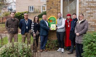 Group of colleagues from Settle, Charlie from the JE3 Foundation, and partners from Astraseal standing next to a newly installed outdoor defibrillator mounted on a brick wall. The defibrillator is bright yellow with a green front panel, and two people in the centre are holding the AED unit.