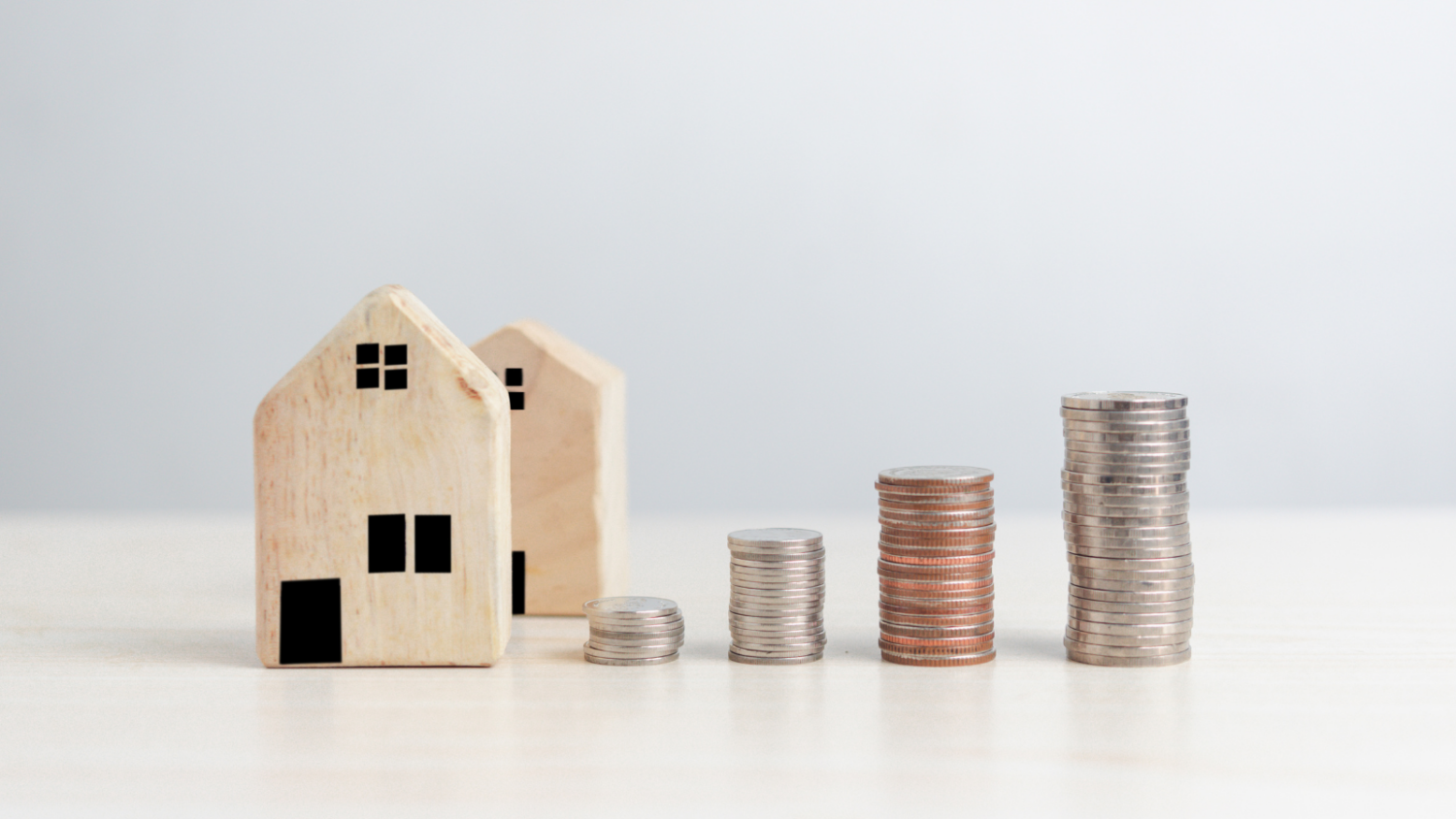 Two small wooden house models placed on a light surface next to four stacks of coins arranged in ascending order, symbolising property investment or increasing house prices.