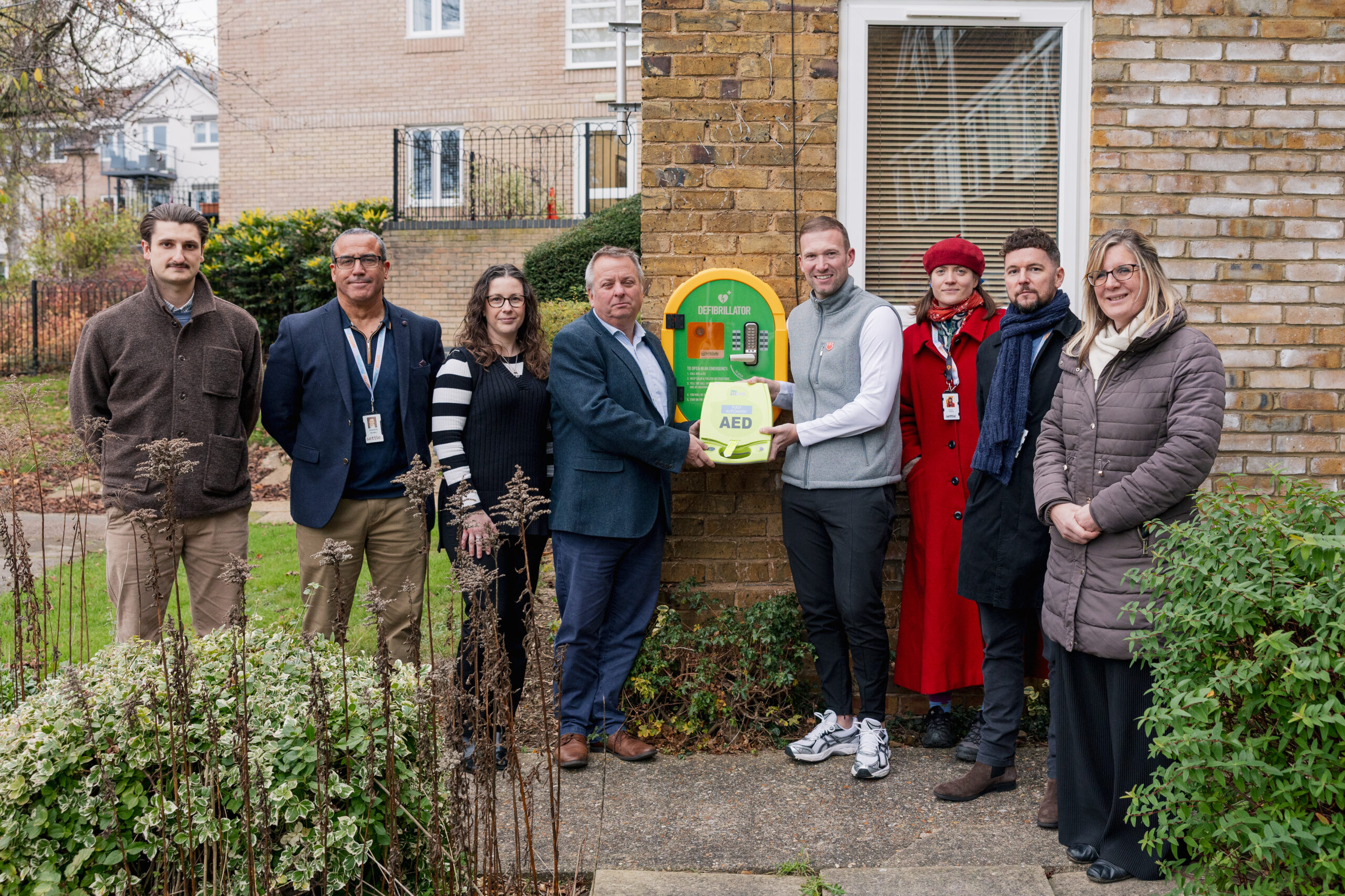 A group of colleagues from Settle, the founder of the JE3 Foundation, and partners from Astraseal standing next to a newly installed outdoor defibrillator mounted on a brick wall in Girton Court, Hitchin.