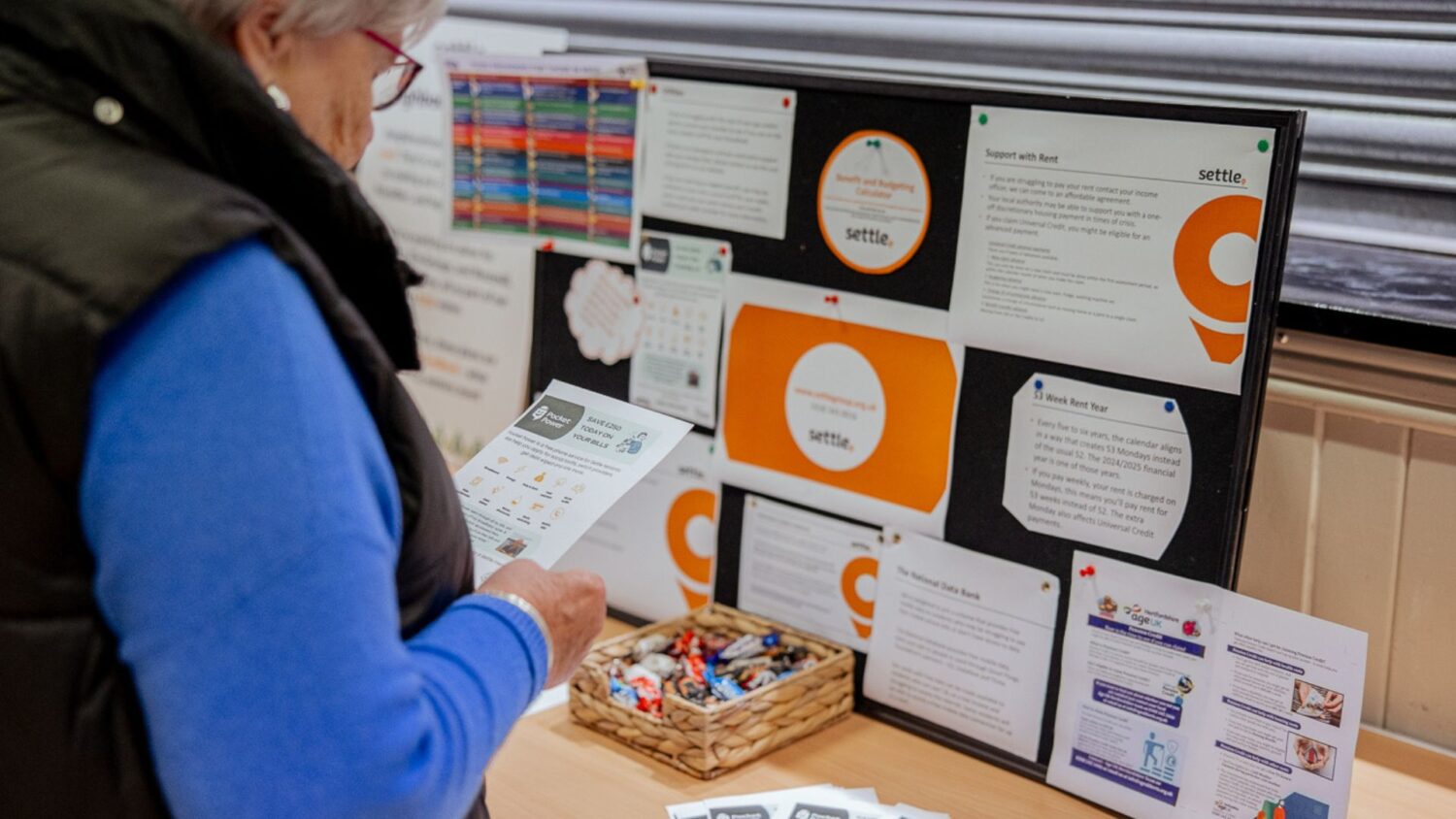 A resident stands at a table reading an information leaflet. In front of them is a display board covered with printed advice sheets and posters from community organisations, including Settle.