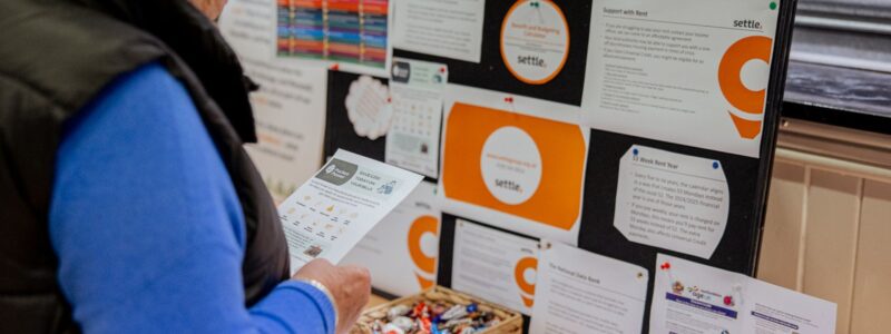 A resident stands at a table reading an information leaflet. In front of them is a display board covered with printed advice sheets and posters from community organisations, including Settle. 