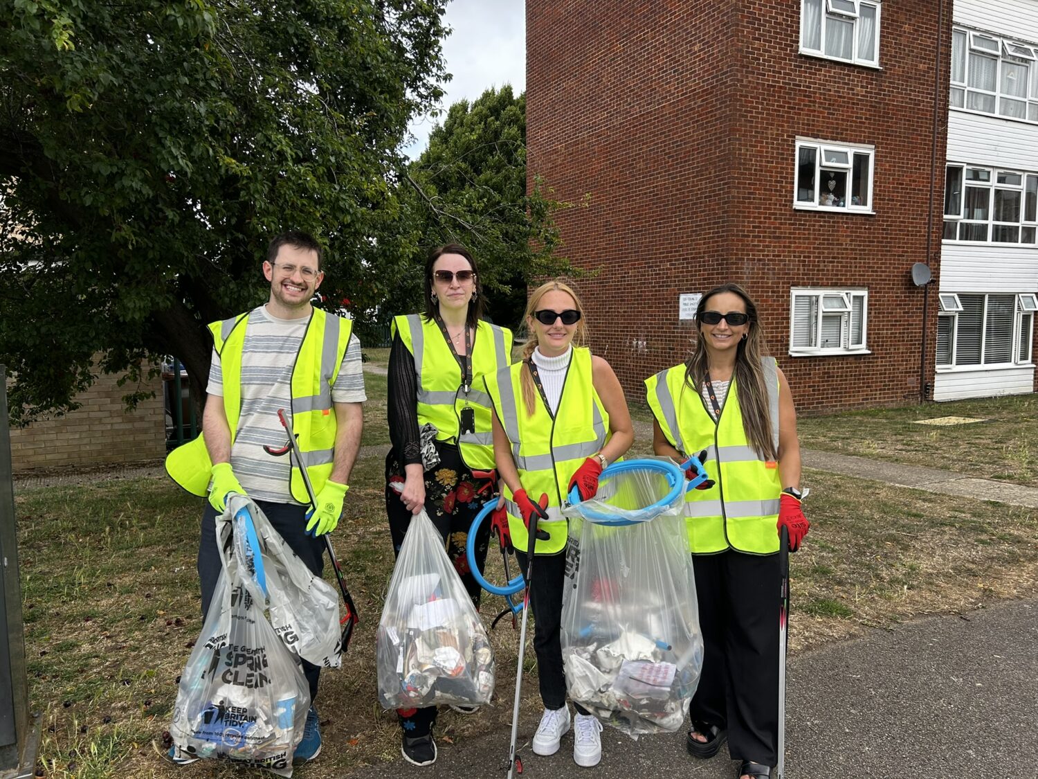 A group of four Settle colleagues wearing high‑visibility vests and gloves stand outdoors beside a residential building, holding litter‑picking tools and large bags filled with collected rubbish. They appear to be participating in a community clean‑up activity, with trees and a grassy area in the background.