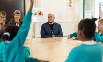 Cllr Sean Prendergast wearing a lanyard, seated at a classroom table with Garden City Academy Pupil Parliament pupils, with pupils raising their hands.