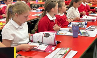 A group of Year 6 pupils in red and white school uniforms sit at a classroom table reading and completing activity booklets. One pupil in the foreground looks down at an open workbook showing an illustration, while others follow along with their copies. The table is covered with books, pencil cases and a water bottle, and the classroom setting includes shelves, materials and bright displays in the background.