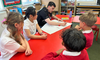 A police officer sits at a red classroom table with a small group of primary school pupils in red and white uniforms, leading a discussion activity. The officer writes notes on a large sheet of paper in the centre of the table while the pupils watch and listen. The surrounding classroom includes shelves of books and stationery, storage folders, and windows letting in natural light.