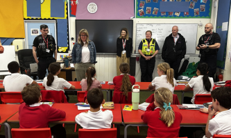 A classroom of primary school pupils in red and white uniforms sit at desks facing the front, where a group of visiting professionals stand and speak. The group includes two police officers in uniform and another person wearing a high-visibility vest, alongside several other adults with ID lanyards. Behind them is a whiteboard with handwritten notes and a large screen, with colourful classroom displays and student photos on the walls.
