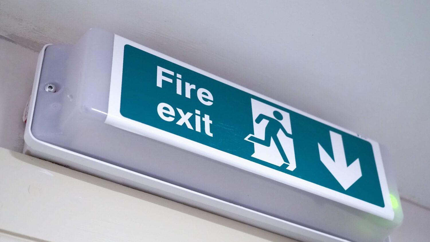 Green and white illuminated fire exit sign mounted high on an interior wall, showing the words “Fire exit” with a running figure symbol and a downward arrow indicating the direction of escape.