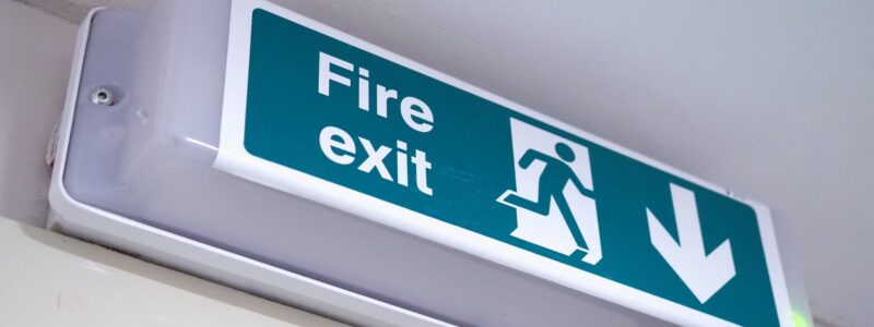Green and white illuminated fire exit sign mounted high on an interior wall, showing the words “Fire exit” with a running figure symbol and a downward arrow indicating the direction of escape.