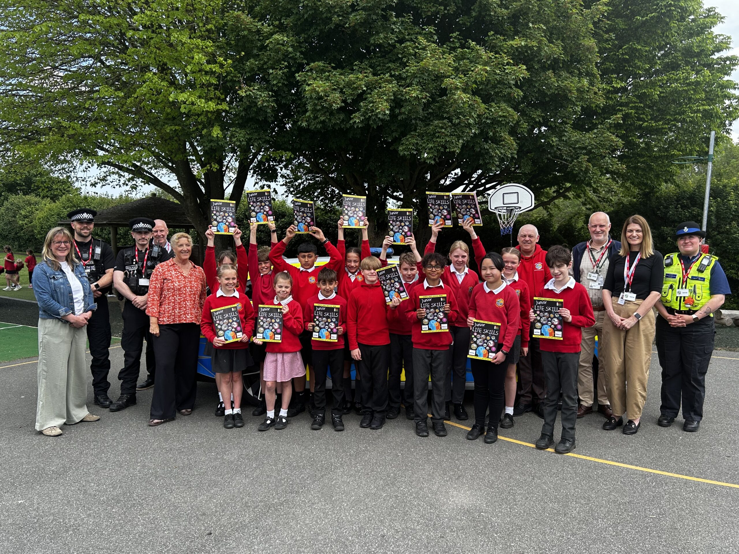 A large group of schoolchildren in red uniforms stand in front of a marked police car in a school playground, holding up “Junior Life Skills” booklets. Several adults stand alongside them, including uniformed police officers and staff wearing lanyards. The setting is outdoors with a basketball hoop, paved play area, and large leafy trees in the background. The children are arranged in rows, some raising the booklets above their heads while others hold them at chest height, and additional children can be seen playing in the distance.