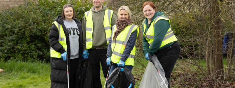 Settle colleagues wearing high-visibility yellow vests stand on a grassy area near bushes and a brick building, holding litter pickers and collection bags. Some of the bags contain rubbish, and all four individuals are wearing gloves as they take part in a community clean-up activity as part of The Great British Spring Clean.