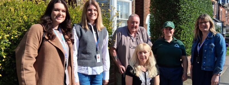 A photo from the Green aiders project launch day. Residents, Settle colleagues and Groundwork East colleagues stand and sit together on a sunlit residential street in front of red brick houses. A resident sits in a wheelchair in the centre foreground, while five others stand nearby. Some individuals wear lanyards and one wears a green uniform-style top and cap. A hedge and building entrances with arched brickwork are visible behind them under a clear blue sky.