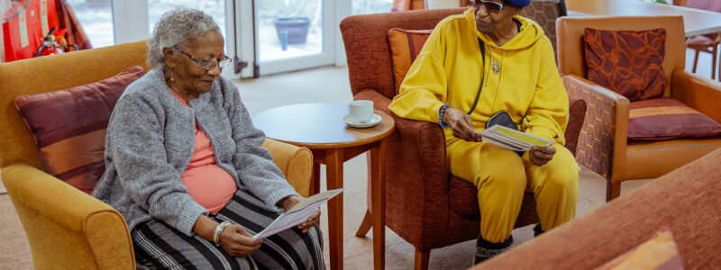 Two residents from an independent living scheme sit in armchairs in a communal lounge area, each holding and reading a leaflet. A small table with a cup sits between them, and large windows with curtains and additional seating are visible in the background.