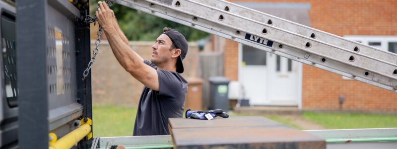 A Settle operative stands next to a work vehicle, securing equipment with a chain. A ladder is positioned overhead, and tools and materials are visible in the back of the vehicle, with residential houses in the background.